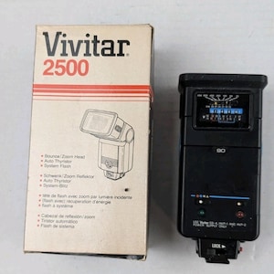 May include: A black Vivitar 2500 camera flash unit positioned next to its original cardboard box. The box features the Vivitar 2500 branding, a line drawing of the flash, and text listing features like Bounce/Zoom Head and Auto Thyristor.