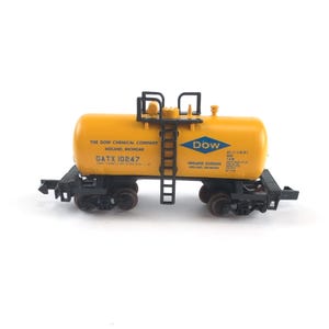 May include: A yellow model tank car with black undercarriage and ladder. The side features blue text reading THE DOW CHEMICAL COMPANY MIDLAND, MICHIGAN, GATX 10247, and a blue Dow diamond logo. Additional technical text is printed on the right side.