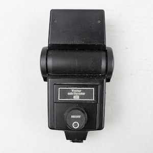 May include: A black Vivitar auto thyristor 283 camera flash unit. The device features a rectangular head, a central control panel with the brand name and model number, and a circular sensor dial positioned below the main display area.