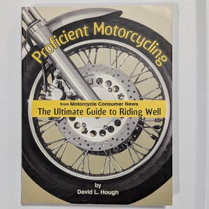 以下が含まれることがあります： デビッド・L・ハフ著『Proficient Motorcycling: The Ultimate Guide to Riding Well』というタイトルのソフトカバー本。表紙には、オートバイの前輪、タイヤ、ブレーキディスク、サスペンションフォークの大きく詳細なグラフィックが白黒で描かれています。