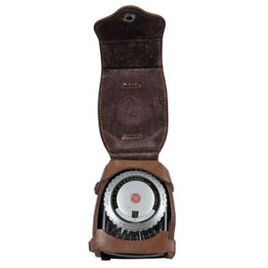 Puede incluir: Un fotómetro vintage en un estuche de cuero marrón. El fotómetro tiene un dial circular con varias marcas y un logotipo rojo y blanco. El estuche está abierto, revelando la esfera del medidor. El cuero tiene un aspecto desgastado.