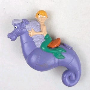 May include: A plastic toy figure featuring a light-skinned character with blonde hair riding a purple seahorse. The character has a green tail and is sitting on an orange saddle. The seahorse has a light purple mane and a curled tail.