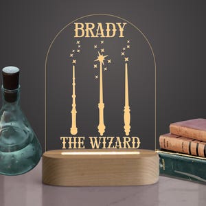 May include: A personalized night light with the name "BRADY" above three wand illustrations and the text "THE WIZARD". The light has a wooden base and a clear acrylic panel. A glass bottle and books are in the background.