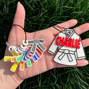 May include: A collection of colorful karate belt keychains and a white karate uniform keychain with the name "Charlie" in red. The keychains are held against a background of green grass.