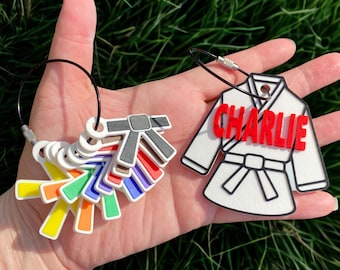 May include: A collection of colorful karate belt keychains and a white karate uniform keychain with the name "Charlie" in red. The keychains are held against a background of green grass.