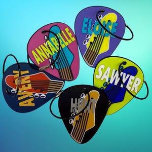 May include: A collection of guitar pick-shaped luggage tags in various colors, each featuring a guitar illustration and a name in bold letters. The tags include names like "Annabelle," "Eloise," and "Sawyer."