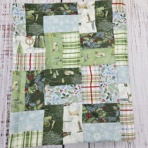May include: A patchwork quilt featuring a variety of winter-themed fabrics. The quilt incorporates squares of green, blue, and white fabrics with patterns of deer, trees, snowflakes, and plaid. The quilt is approximately 30 inches by 30 inches.