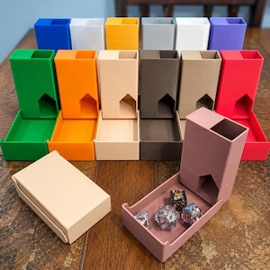 Puede incluir: Una colección de torres y bandejas para dados de colores, incluyendo verde, naranja, beige, marrón y rojo. Una torre rosa está abierta, mostrando varios dados. Las torres están dispuestas sobre una superficie de madera.