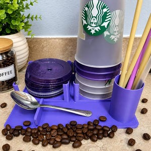 Può includere: Un organizer viola per tazze da caffè con un cucchiaio, coperchi e cannucce. L'organizer contiene diverse tazze trasparenti con un logo verde e bianco. I chicchi di caffè sono sparsi intorno all'organizer.