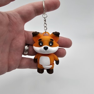 Puede incluir: Un llavero de zorro naranja con vientre y cara blancos, orejas y pies marrones y ojos negros. El zorro está sujeto a una cadena y un llavero plateados. Un pequeño accesorio decorativo.