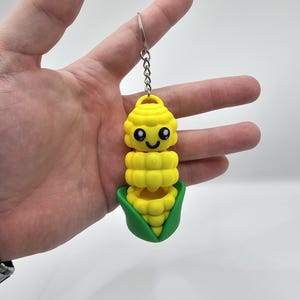 Puede incluir: Un llavero con forma de maíz amarillo con una cara sonriente, una cáscara verde y un llavero plateado. El maíz tiene un diseño segmentado y se sostiene en una mano sobre un fondo blanco. Un divertido accesorio temático de comida.