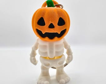 Puede incluir: Un llavero impreso en 3D que muestra una figura de esqueleto blanco con una cabeza de calabaza de color naranja brillante. La calabaza tiene un tallo verde y ojos, nariz y boca negros recortados. Un anillo de metal plateado está unido a la parte superior.
