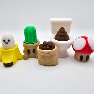 Pode incluir: Uma coleção de figuras caprichosas em estilo cartoon. Inclui uma banana sorridente, um cacto num vaso, um saco de grãos de café, uma sanita com um emoji de cocó e um cogumelo vermelho e branco. As figuras são coloridas e têm um design divertido.