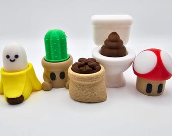 Könnte beinhalten: Eine Sammlung skurriler, cartoonartiger Figuren. Enthält eine lächelnde Banane, einen Kaktus im Topf, einen Sack Kaffeebohnen, eine Toilette mit einem Kackhaufen-Emoji und einen rot-weißen Pilz. Die Figuren sind farbenfroh und haben ein verspieltes Design.