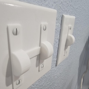 Könnte beinhalten: Zwei weiße Lichtschalterabdeckungen sind an einer hellblauen Wand montiert. Die größere Abdeckung hat einen horizontalen Kippschalter mit abgerundeten Enden. Die kleinere Abdeckung hat einen einzelnen Kippschalter mit abgerundetem Ende.