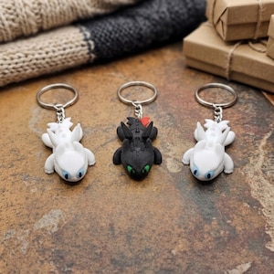 May include: Three dragon-shaped keychains. Two are white with blue eyes, and one is black with green eyes. Each keychain has a silver ring. The keychains are on a textured brown surface.