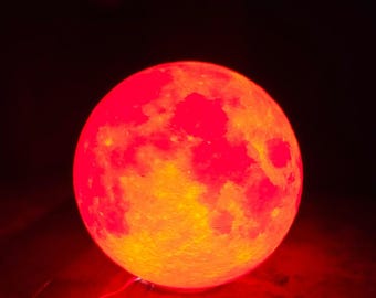 May include: A glowing, spherical lamp resembling a red and orange moon. The lamp has a textured surface with a crater-like design, and is lit from within, casting a warm, reddish glow against a dark background.