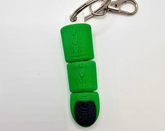 May include: A green keychain shaped like a finger with a black fingernail. The finger has a textured surface resembling skin folds and is attached to a silver metal split ring and a lobster claw clasp. The top of the finger has a white connector.