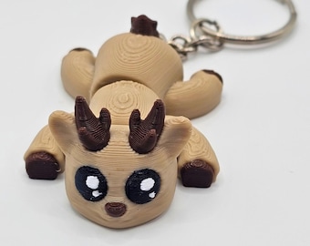 May include: A 3D-printed tan deer keychain featuring dark brown antlers, hooves, and a small tail. The deer has large black eyes with white highlights and a small brown nose. The figure is attached to a circular silver metal split ring.