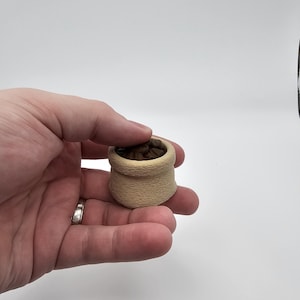 Può includere: Un piccolo cestino in miniatura beige pieno di oggetti marroni, possibilmente chicchi di caffè o simili. Il cestino ha un aspetto strutturato ed è tenuto in una mano su sfondo bianco. Il cestino è alto circa 3,8 cm.