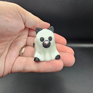 May include: A 3D-printed figurine of a cat in a ghost costume. The cat is white with black ears, eyes, and paws, and a pink nose. The figurine is held in a hand against a dark background.