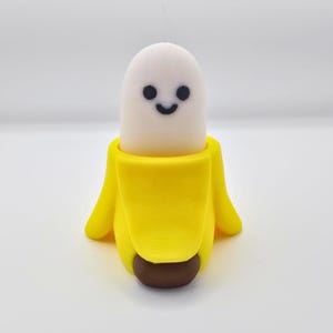 May include: A whimsical, handcrafted banana-shaped figurine. The bright yellow peel cradles a smiling, white, oblong figure with black dot eyes and a curved mouth. The base is a brown, rounded shape. The overall design is playful and cartoonish.