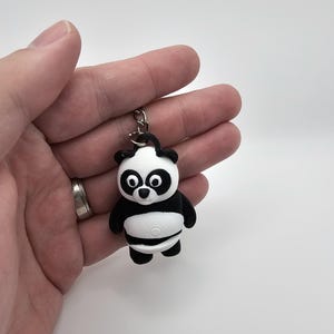 Peut inclure: Un porte-clés panda noir et blanc. Le panda a une tête et un ventre blancs, avec des détails noirs sur les oreilles, les bras et les jambes. Le porte-clés est attaché à un anneau en métal argenté.