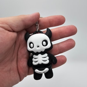 Puede incluir: Un llavero con temática de esqueleto en blanco y negro. El llavero presenta una figura de dibujos animados con una cabeza en forma de calavera, cuernos y un diseño de caja torácica. La figura está unida a un llavero de color plateado.