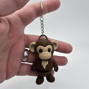 Può includere: Un portachiavi a forma di scimmia marrone con viso, orecchie e petto color beige chiaro. La scimmia ha occhi neri e un anello portachiavi argentato. Il portachiavi è realizzato in plastica dura.