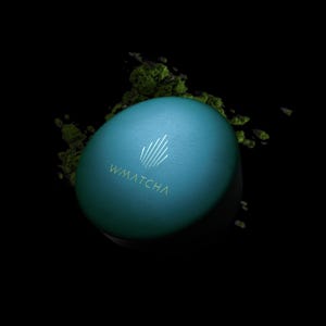 May include: A round teal container with a matte finish, featuring the white text WMATCHA and a stylized leaf icon on the lid. The container is surrounded by scattered green matcha powder against a solid black background.