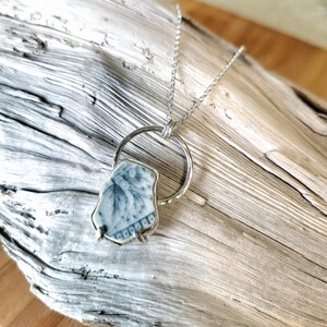 May include: A silver chain necklace with a blue and white ceramic pendant. The pendant is a piece of broken china with a floral design. The pendant is suspended from a silver ring.