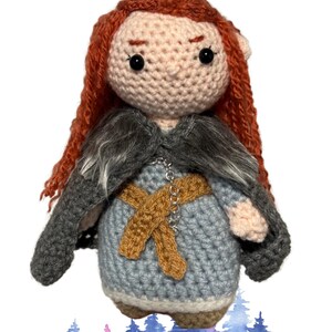 May include: Crocheted doll with long red hair, wearing a grey fur-trimmed cloak, a blue dress with a brown belt, and a silver chain.