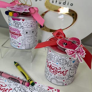 Puede incluir: Dos recipientes de regalo con temática de San Valentín. Cada recipiente blanco presenta un dibujo lineal negro y el texto "Happy Valentine's Day!". Uno tiene un diseño de robot, el otro un corazón. Ambos tienen crayones y cintas rosas.