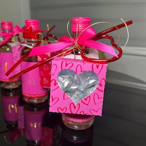 May include: A collection of small bottles with pink labels and caps, adorned with pink ribbons and heart-shaped decorations. A pink box with a heart-shaped cutout reveals silver-wrapped candies. The overall theme is Valentine's Day.