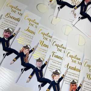 Puede incluir: Colgadores de puerta blancos con texto dorado e ilustraciones de temática marinera. Cada colgador presenta la cara de una persona y las palabras "Capitan de Me" y un nombre. El texto incluye frases como "Eres el encargado de hacer trabajo mas divertido."