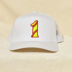 Op de afbeelding: Witte truckerpet met een mesh achterkant en een gebogen klep. De pet heeft een groot, geborduurd nummer "1" in geel met rode stiksels, dat op een softbal lijkt.