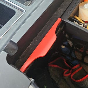 May include: A car's center console, open to reveal various items. A water bottle sits in a cup holder. Inside the console are tape, tools, a small orange ball, and a pair of gray and red gloves. A red plastic divider is visible.