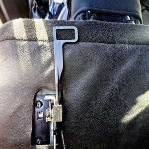 May include: A black metal car seat latch release tool. The tool is inserted into the car seat latch mechanism. The image shows the tool in use, with the latch partially released. The background is a dark gray car seat.