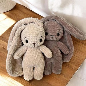 May include: Two handmade crochet bunny dolls. One is light beige, the other is a muted mauve. Both have long floppy ears, black eyes, and a stitched nose and mouth. The bunnies are sitting on a wooden surface.