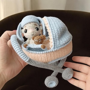 Puede incluir: Una muñeca de bebé hecha a mano en ganchillo en un cochecito azul y melocotón. La muñeca lleva un gorro azul y sostiene un pequeño osito de peluche. El cochecito tiene ruedas grises y está sostenido en las manos de una persona. La cara de la muñeca tiene ojos negros y una pequeña sonrisa.