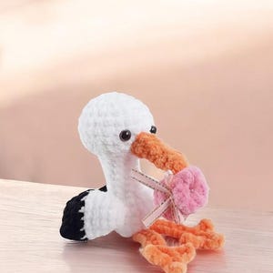 Puede incluir: Un juguete de cigüeña de ganchillo hecho a mano en blanco, negro y naranja, que sostiene un paquete rosa atado con una cinta. La cigüeña tiene ojos negros y un pico y patas naranjas. El juguete está hecho de hilo suave.