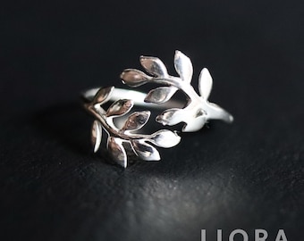May include: A silver-colored ring featuring a delicate leaf design. The ring has two branches of leaves that curve around the finger. The leaves have detailed veining, and the ring is set against a dark background.