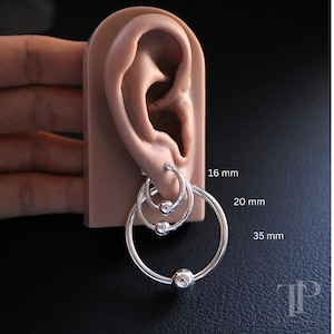 Op de afbeelding: Drie zilveren oorringen met kogelaccenten, maten 16 mm, 20 mm en 35 mm, getoond op een huidkleurig oor model. De oorbellen zijn cirkelvormig en hebben een gepolijste afwerking, tegen een donkere achtergrond.