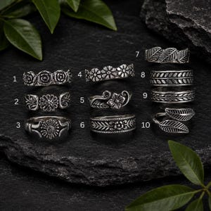 Könnte beinhalten: Eine Sammlung von zehn silberfarbenen Metallringen, die in zwei Spalten angeordnet und von eins bis zehn nummeriert sind. Jeder Ring weist ausgeprägte geprägte Pflanzen- oder Blumenmuster auf, darunter Rosen, Gänseblümchen, Blätter und Ranken, mit einem oxidierten Finish.