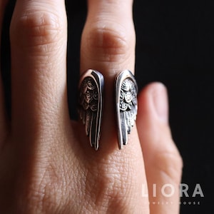 Puede incluir: Un anillo plateado con un dise&ntilde;o abierto, con motivos de alas detallados en cada lado. El anillo se lleva en un dedo, mostrando los intrincados detalles de las plumas. La joyer&iacute;a es de LIORA JEWELRY HOUSE.