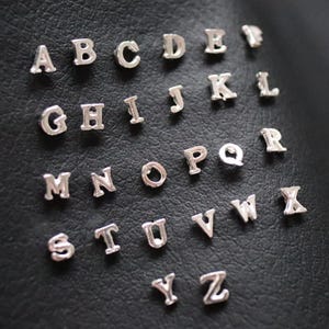 Könnte beinhalten: Silberne Buchstaben-Anhänger auf schwarzem Hintergrund. Die Buchstaben in einer Serifenschrift sind verstreut. Die kleinen, glänzenden Buchstaben reflektieren das Licht. Ideal für Schmuckherstellung oder Bastelprojekte.