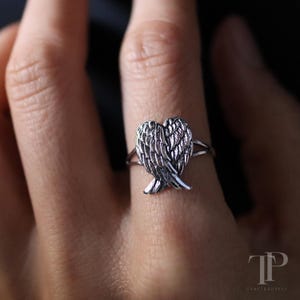 May include: A silver ring featuring a detailed wing design. The ring's focal point is a pair of stylized wings, with intricate feather detailing. The ring is shown on a finger, highlighting the delicate craftsmanship and design.