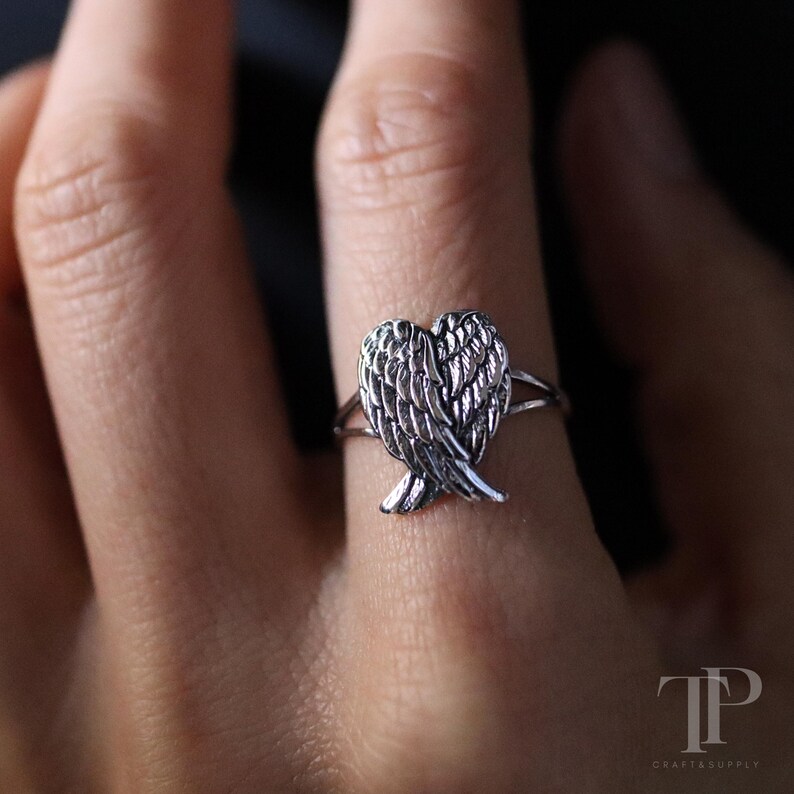 May include: A silver ring featuring a detailed wing design. The ring's focal point is a pair of stylized wings, with intricate feather detailing. The ring is shown on a finger, highlighting the delicate craftsmanship and design.