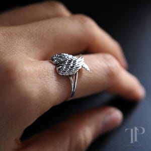 May include: A silver-colored ring featuring a detailed wing design. The ring's focal point is a sculpted wing motif, with intricate feather detailing. The ring is shown on a finger, highlighting the design's delicate craftsmanship and the ring's overall aesthetic.