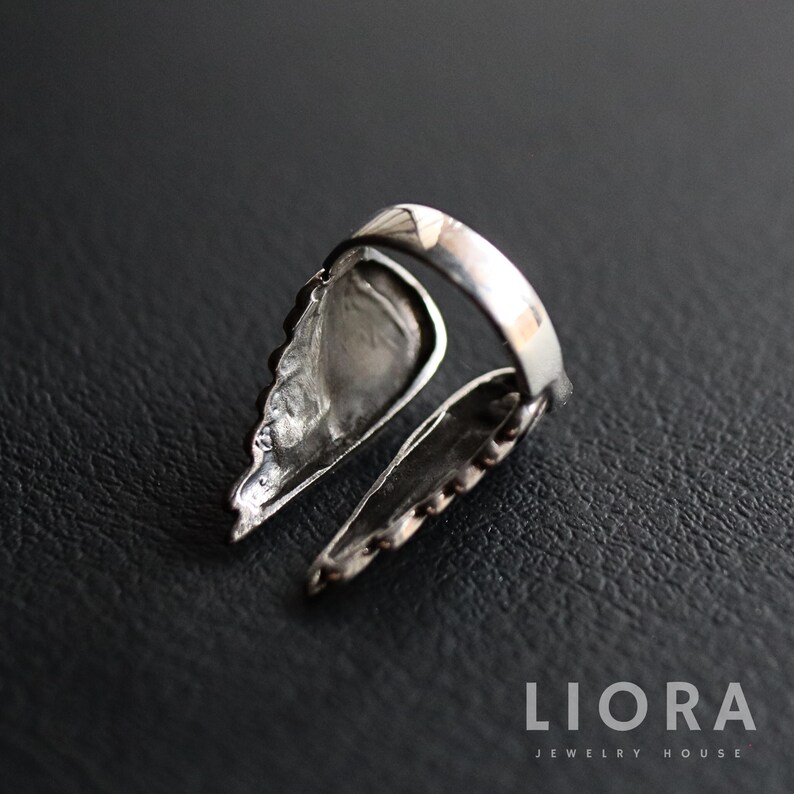 Puede incluir: Un anillo plateado con dise&ntilde;o de ala. El anillo presenta dos alas estilizadas que se extienden desde la banda, con texturas detalladas en forma de plumas. El anillo est&aacute; sobre un fondo oscuro.